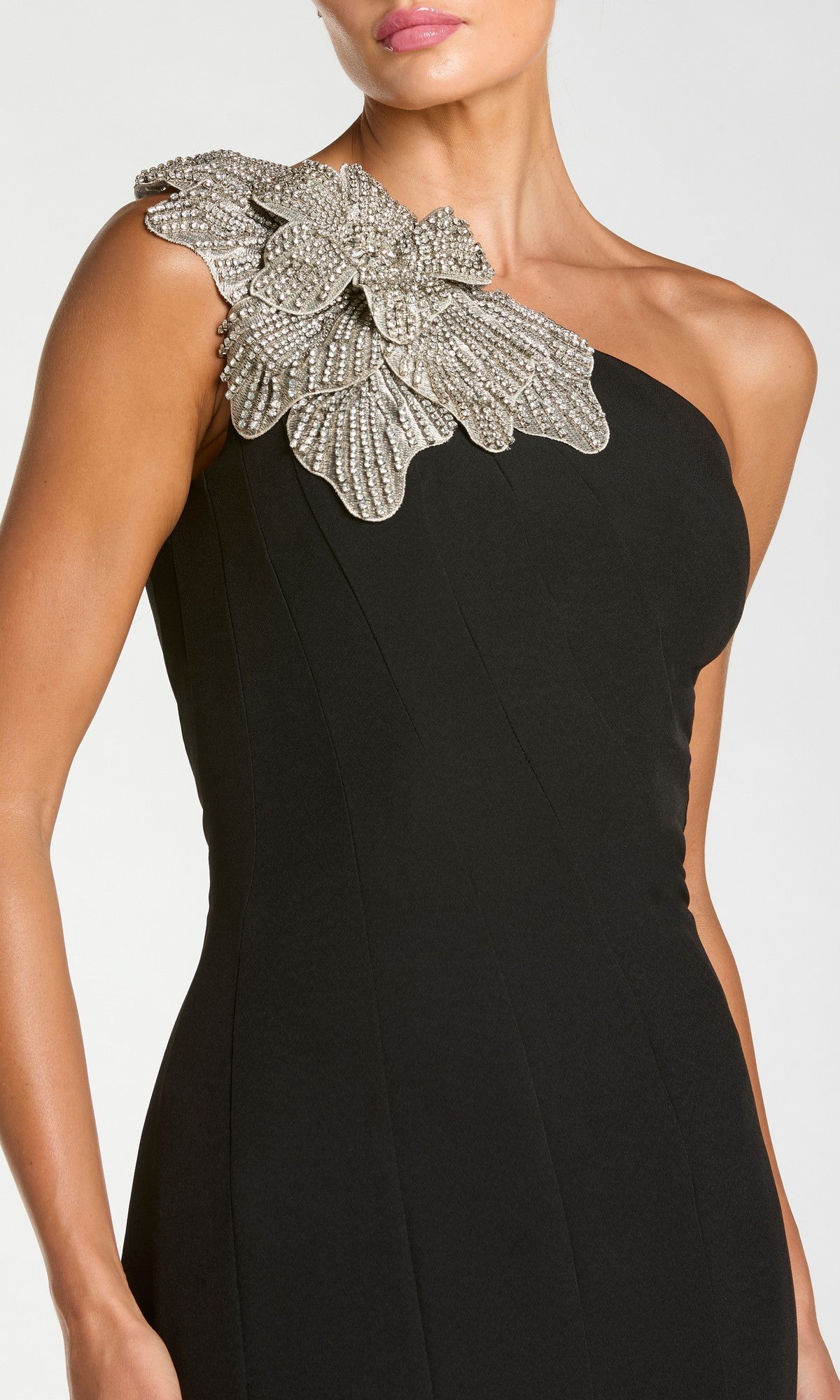 A woman wears the Mac Duggal 13221 Tea-Length Party Dress, a black crepe one-shoulder design adorned with an eye-catching embellished floral applique featuring large beaded detailing on the strap.